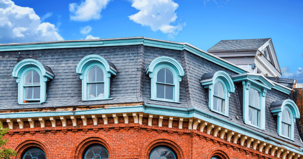 A view of a grey and brick colored house with mansard roofinf and windows under the blue sky with clouds