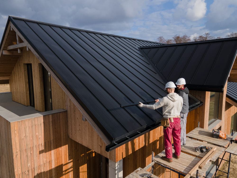 A view of two construction workers placing a metal roof on top of a house standing on wooden panels