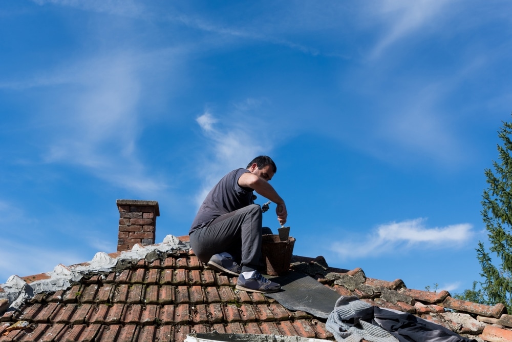 The man sitting on the roof of an old house and repairing tile with cement in the village on a sunny day.