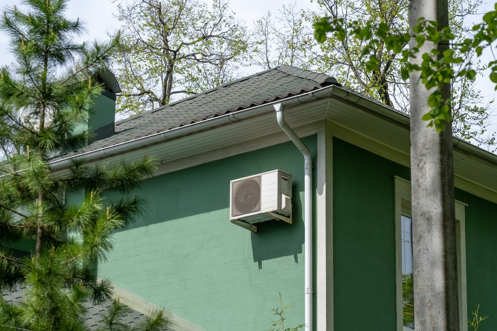 Green stucco house with roof, chimney and rain gutter with downspout, soffit board, outdoor air conditioner unit among trees 